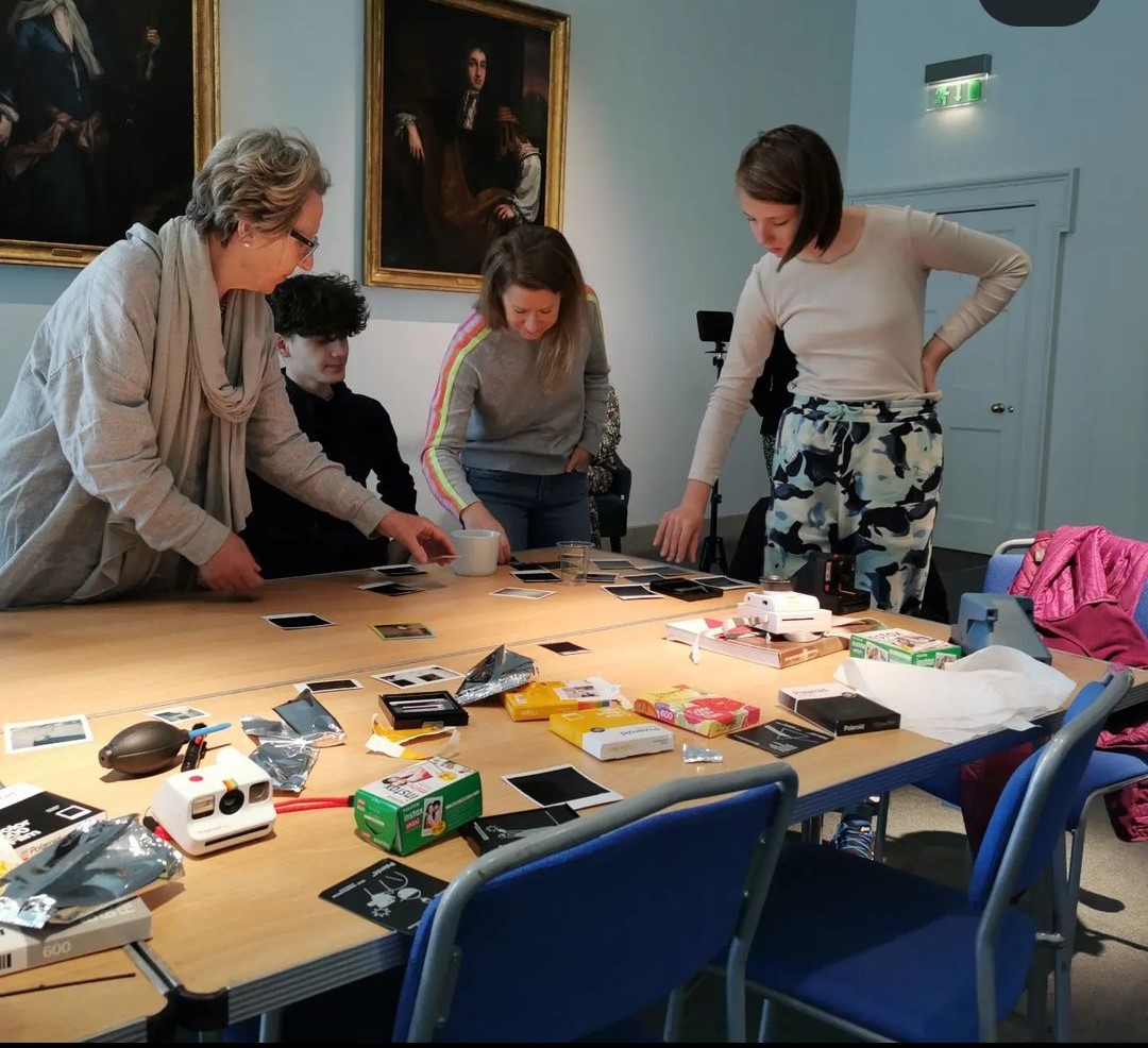 A photograph of the group, looking at the polaroid they have taken. They have placed the photos on the table, and are looing down at them.